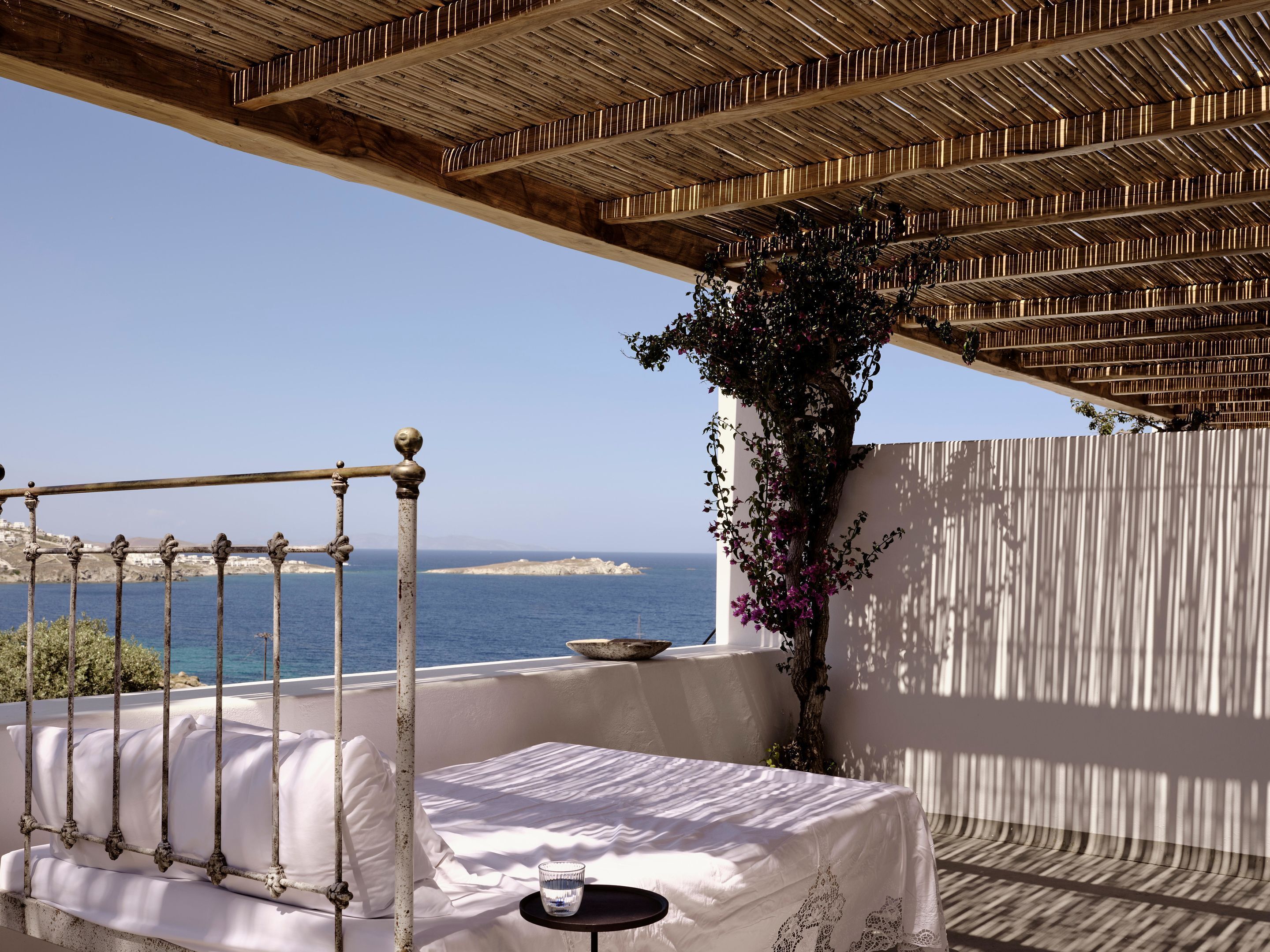 A balcony with a view of the sea, a table with a glass, and a bed with white bedding.