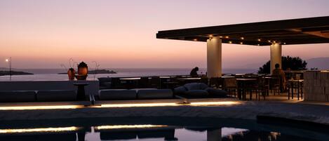 Zona junto a la piscina con asientos y vista al océano al atardecer.