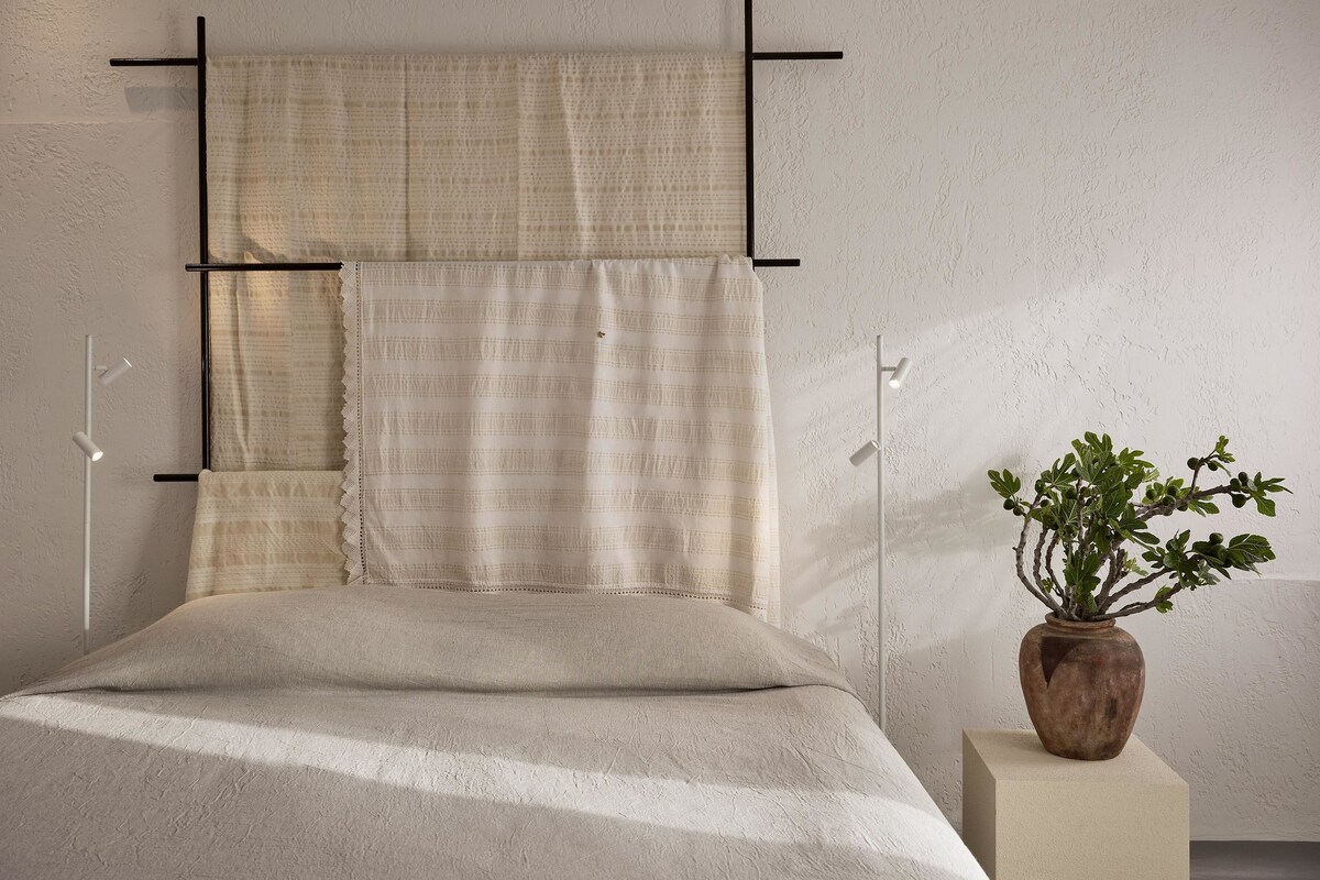 Ein Bett mit beiger Bettdecke und zwei Kissen, eine Zimmerpflanze in einem Holztopf und eine weiße Wand mit strukturierter Oberfläche.