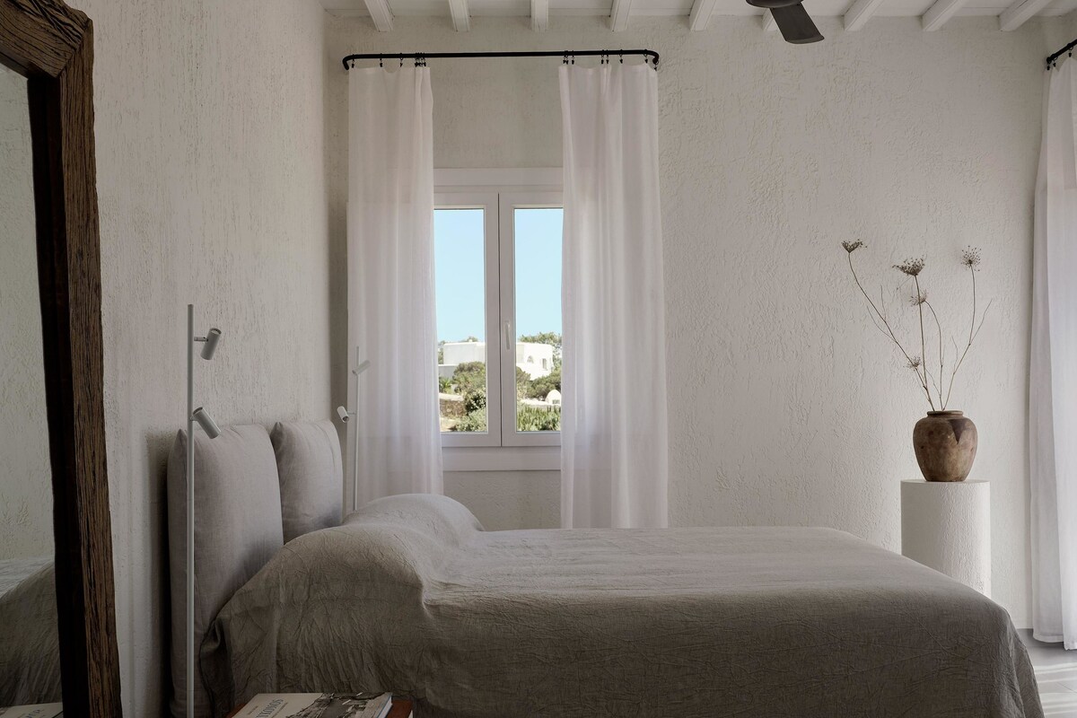 Ein Schlafzimmer mit einem Bett, einem Nachttisch mit Büchern, einem Fenster mit Vorhängen und einer Vase mit getrockneten Blumen.