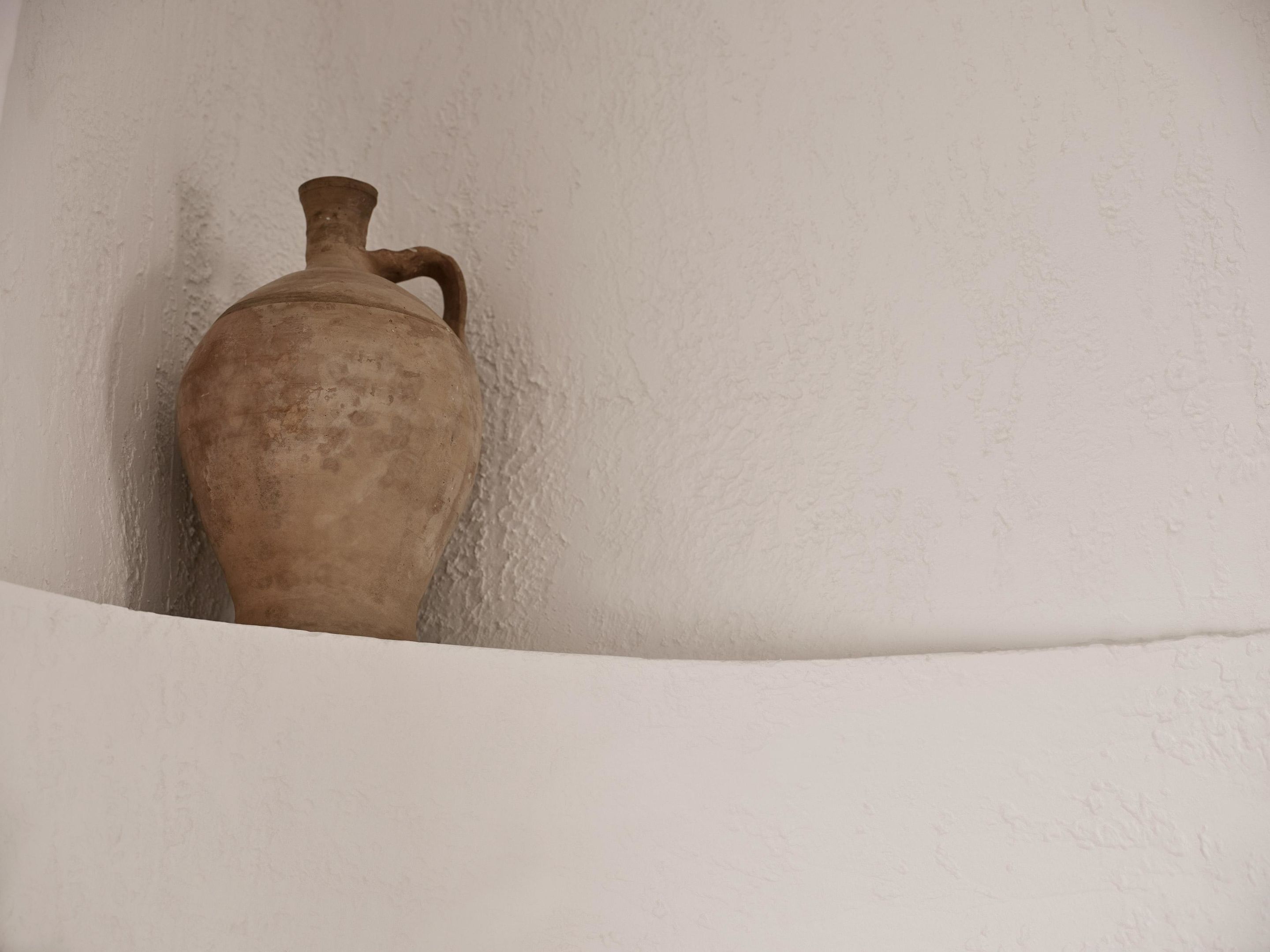 Una jarra de cerámica beige, grande y de un solo cuerpo, apoyada contra una pared blanca con textura.