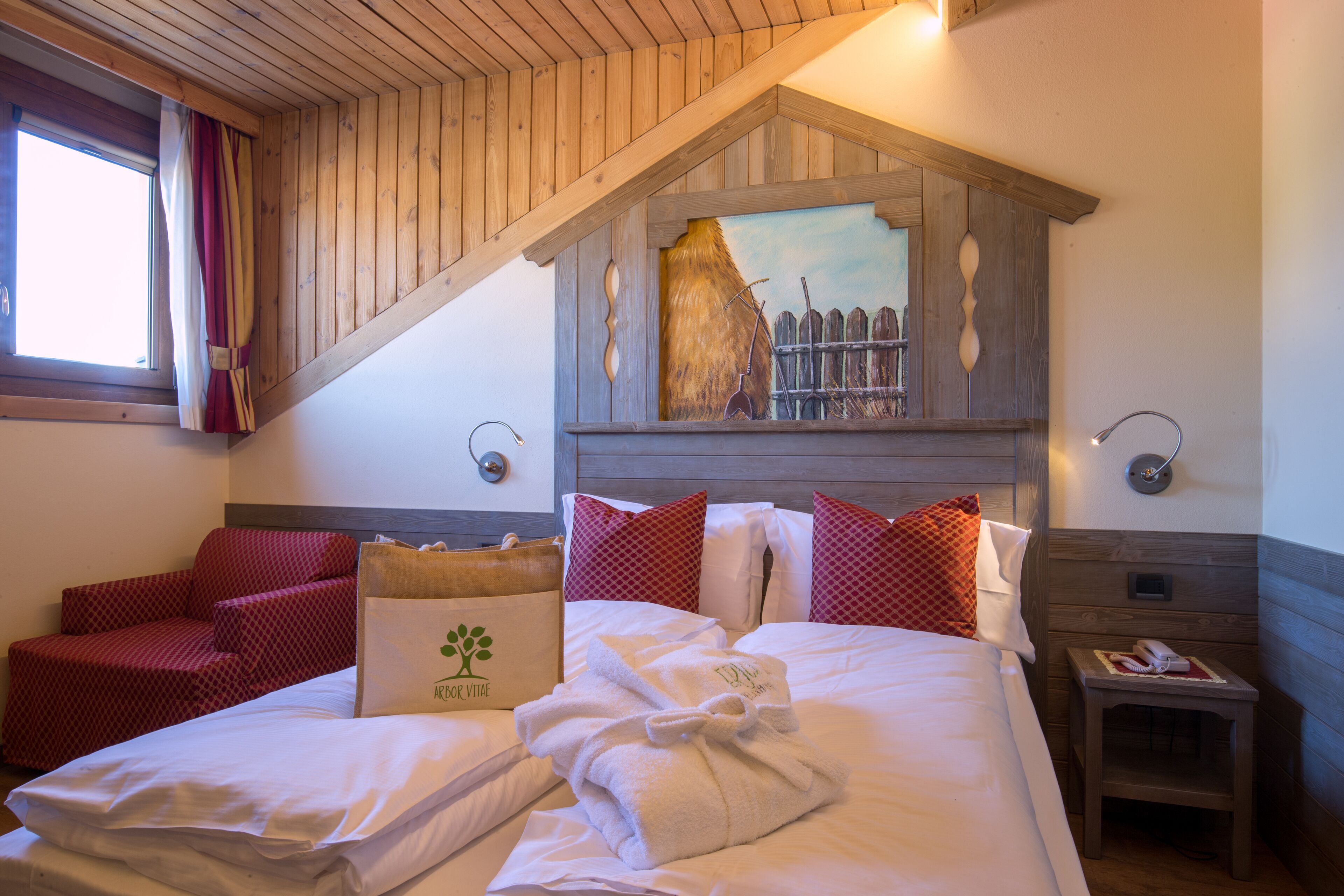 Ein Hotelzimmer mit Holzboden, einem Bild einer Ziegelmauer, einem Bett mit weißen Bettwäsche, roten und weißen Kissen, einem kleinen Nachttisch mit Lampe und einem roten Sessel.