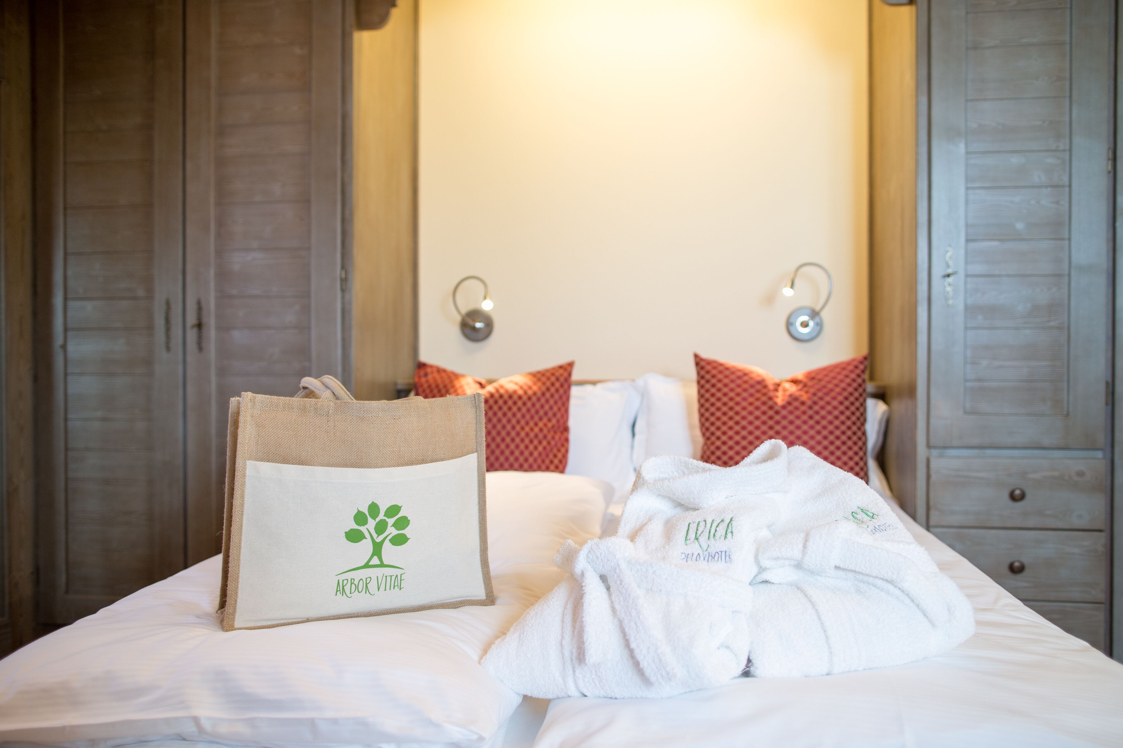 A neatly made bed with a white bathrobe and a burlap bag with a logo, set against a wall with two sconces and patterned pillows.