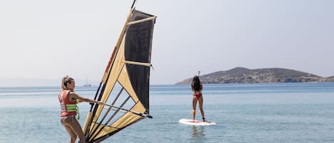Dos personas practican windsurf en un mar tranquilo, bajo un cielo despejado, con tierra a lo lejos.