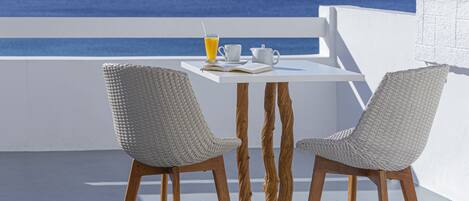 Una mesa blanca con dos sillas de mimbre, un vaso de jugo de naranja, una taza blanca y un libro en un balcón con vistas al mar.
