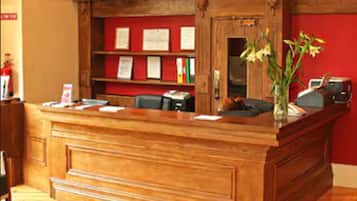 Un mostrador de recepción de madera con una pared roja de fondo, una estantería con diplomas y un florero con flores sobre el mostrador.