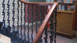 Una escalera con barandillas negras ornamentadas y un pasamanos de madera, que conduce a una habitación con una estantería y una lámpara.
