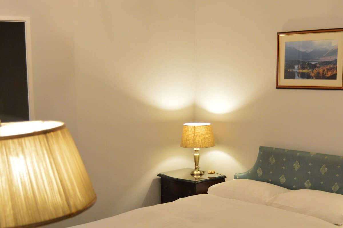 A hotel room with a bed, a desk with a lamp, a nightstand, and a framed picture on the wall.