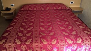 A bed with a red and gold patterned bedspread, wooden headboard, and bedside tables with electronic devices. - Hotel Nettuno (Milan)