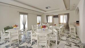 A spacious dining area with white tables and chairs, a flat-screen TV mounted on the wall, and a potted plant. - Soula Naxos (Naxos)