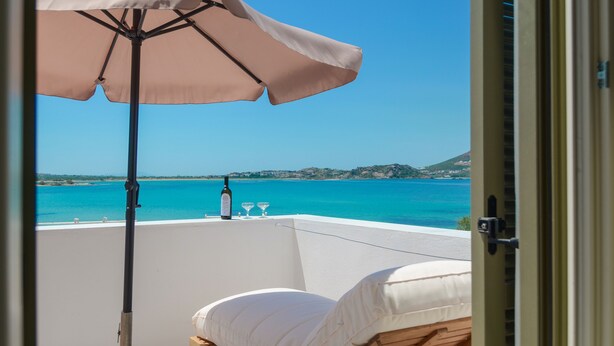A balcony with a beige umbrella, a bottle, and two glasses on a white table. - Soula Naxos (Naxos)