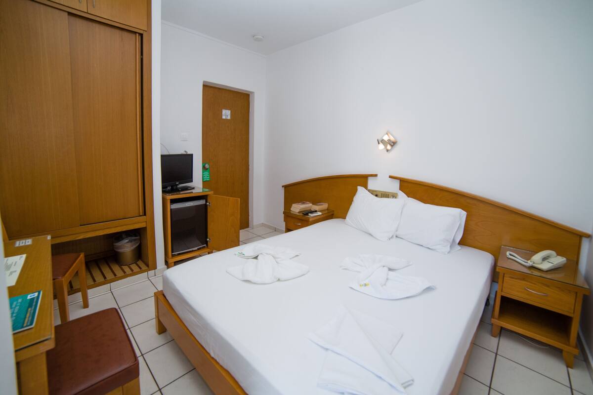 A hotel room with a bed, a wooden wardrobe, a desk with a computer, a telephone, and a small table with a lamp.