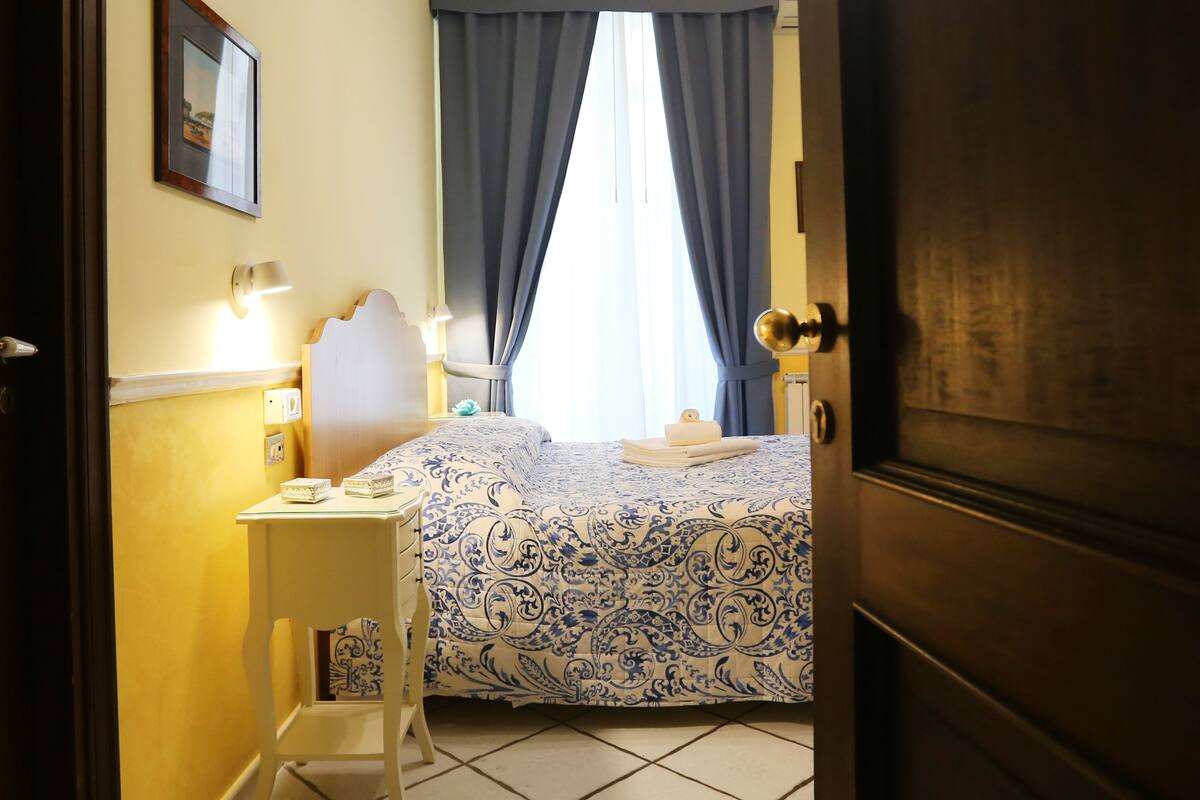Habitación de hotel con cama, mesita de noche y una silla. Tiene una ventana con cortinas, un cuadro en la pared y una puerta.