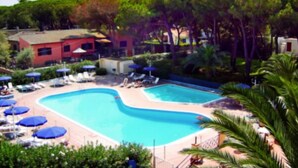 Piscina con sillas de descanso y sombrillas, rodeada de vegetación y edificios.