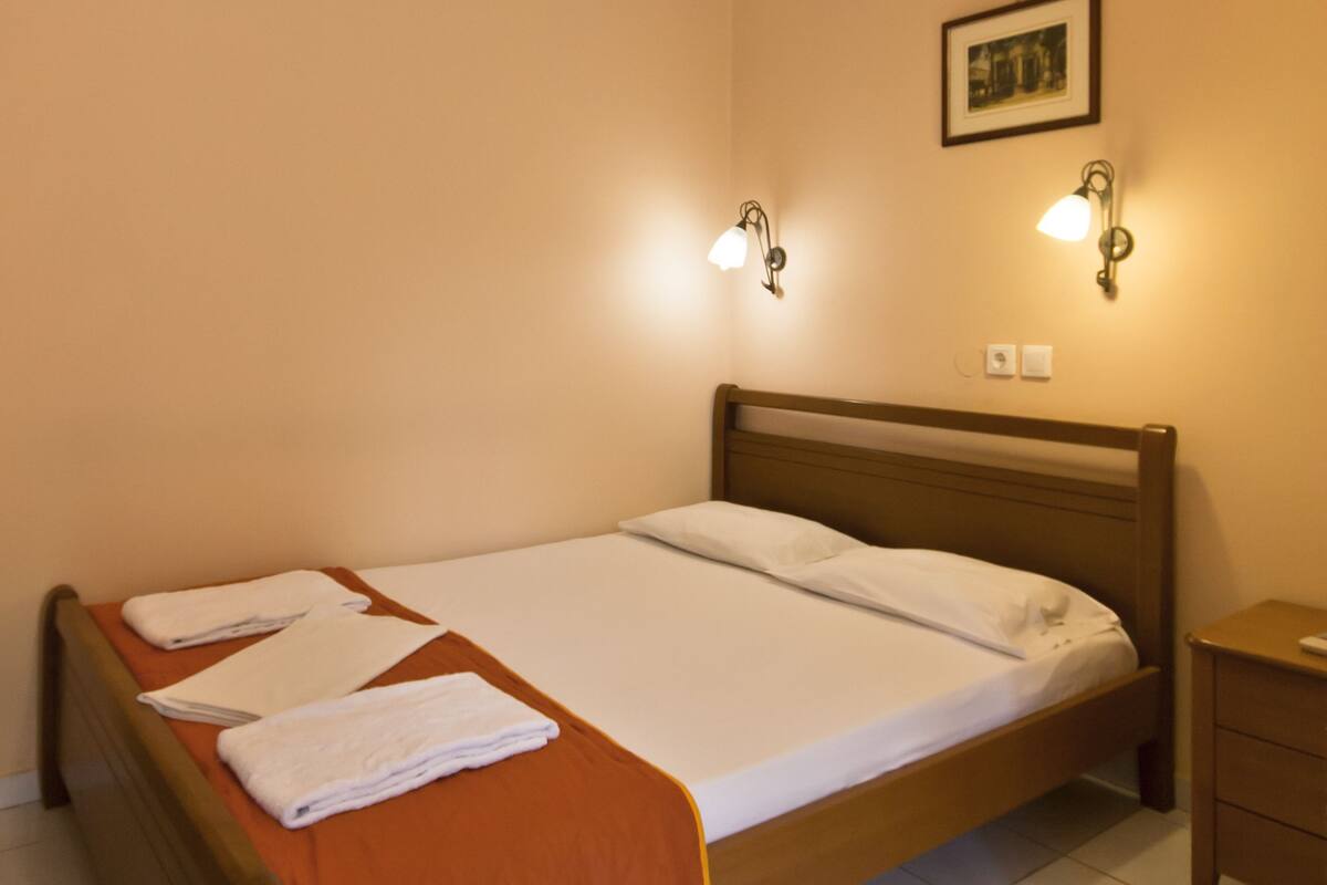 Un lit simple avec une tête de lit en bois, une couvre-lit marron et du linge de lit blanc. Deux appliques murales sont fixées de chaque côté du lit.