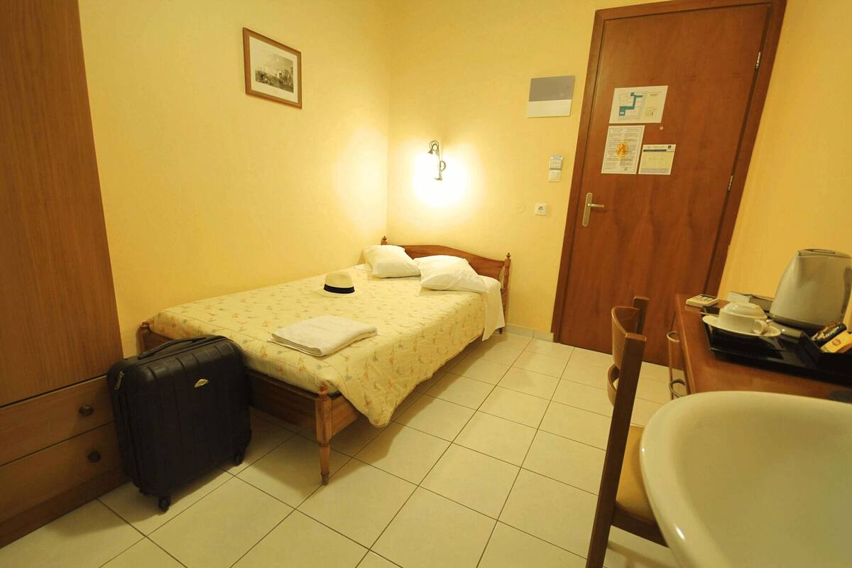 Une petite chambre à coucher, bien rangée, avec un lit simple, une table de chevet avec une lampe, une chaise et une valise.