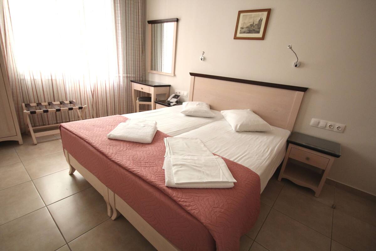 Un lit double bien fait, avec du linge de lit blanc, une table de chevet, un petit bureau avec un téléphone et une fenêtre avec des rideaux.
