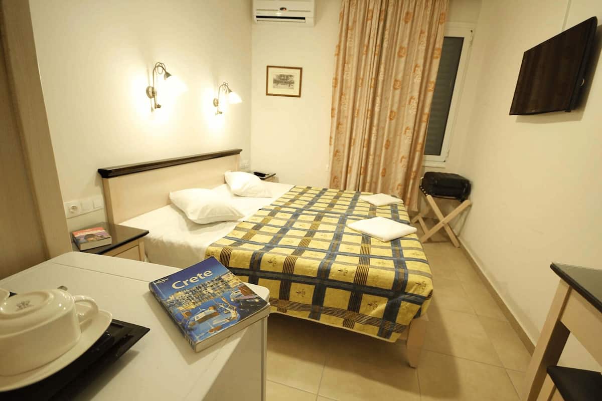 Une petite chambre d’hôtel avec un lit, une table de chevet, une chaise, un bureau et une fenêtre avec des rideaux.