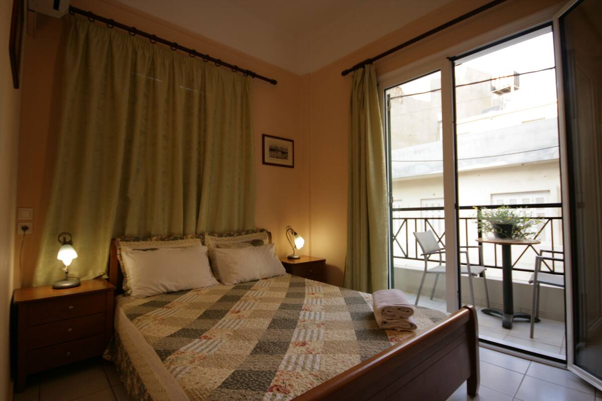 Une chambre à coucher comprenant un lit, des tables de chevet, une fenêtre avec des rideaux et un balcon avec une table et des chaises.