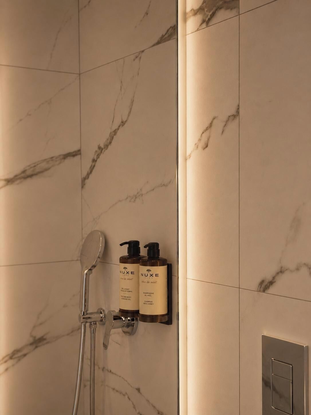 A bathroom with marble walls, a shower, and two Nuxe shampoo bottles.