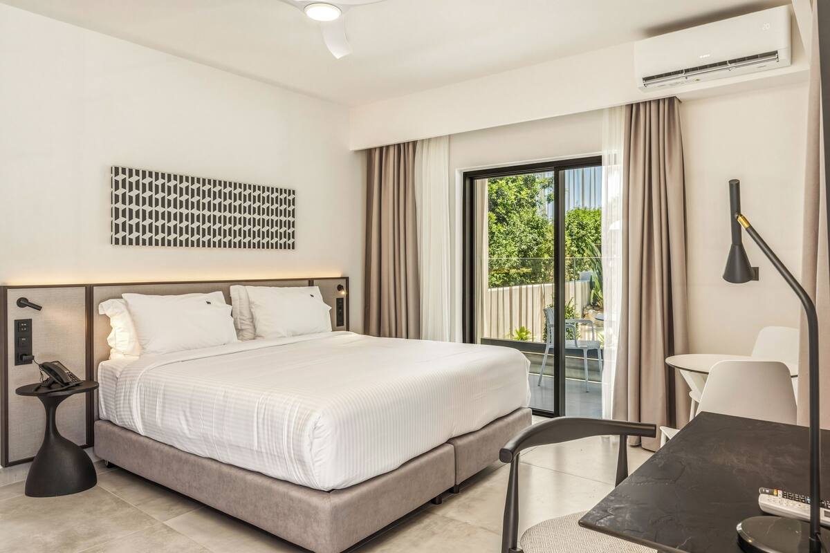 A modern hotel room with a bed, a desk, a chair, a phone, an air conditioner, and a view of greenery outside.