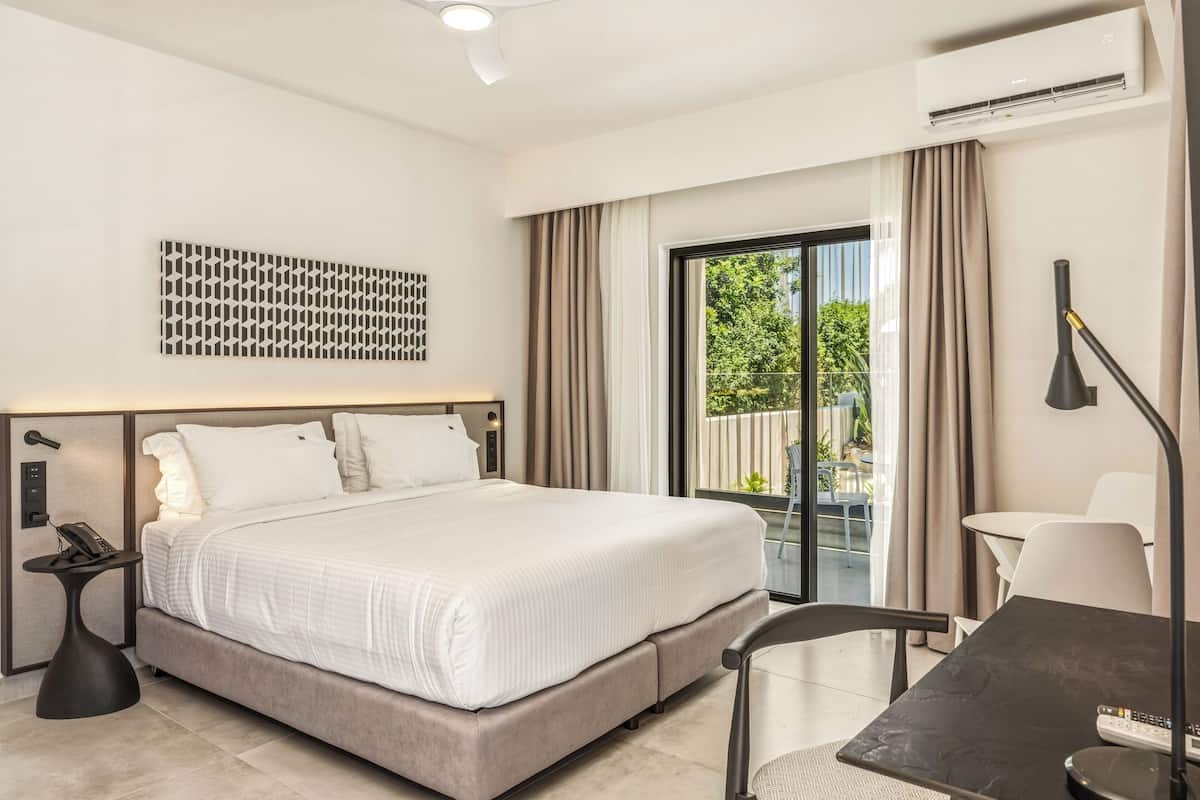 A modern hotel room with a bed, a desk, a chair, a phone, an air conditioner, and a view of greenery outside.