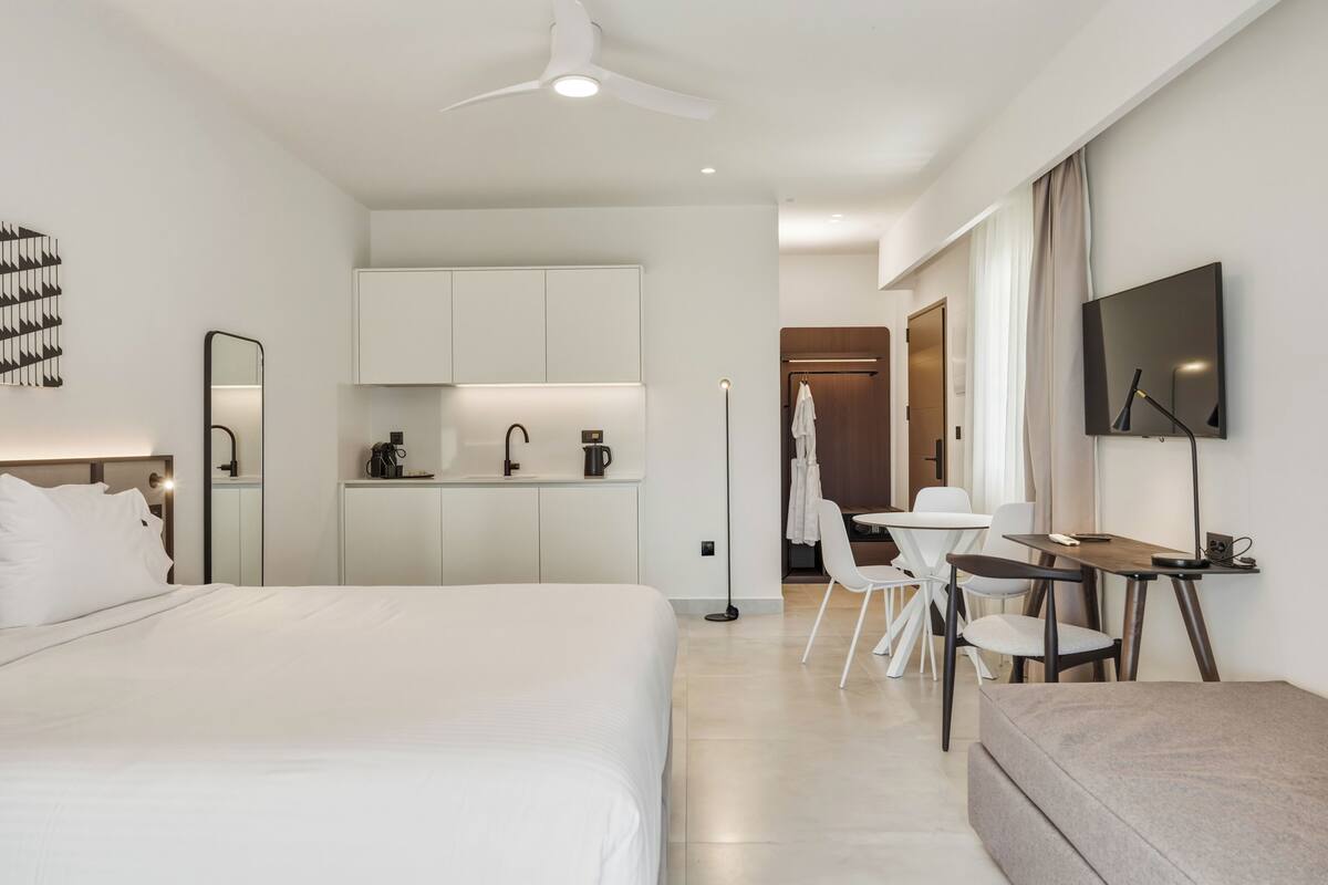A modern hotel room with a large bed, a desk, a chair, a small kitchenette, and a TV.