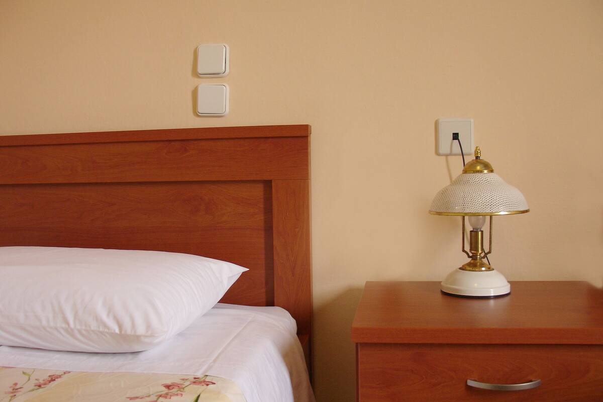 A neatly made bed with white linens and a floral-patterned blanket, a wooden nightstand with a lamp, and a wall socket.