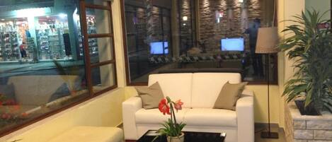 A living room with a white sofa, a black coffee table with a plant, and a large window with a view of a store and a building with multiple screens.
