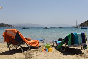Una playa con dos sillas de playa, un cubo verde y un juguete. Se ven barcos en el agua.
