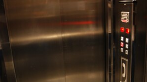 Een lift met een metalen afwerking en een bedieningspaneel met noodknoppen en een telefoon.