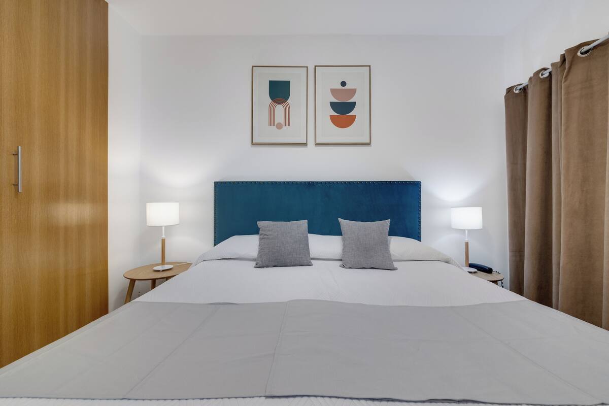 A neatly made bed with a blue headboard, two bedside tables with lamps, and two framed artworks on the wall.