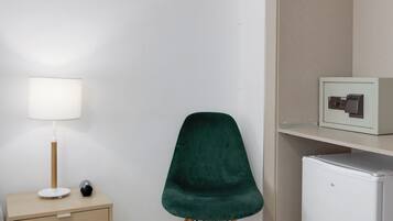 Una habitación con una silla verde, una mesita de noche con una lámpara, un armario blanco pequeño y un refrigerador mini blanco.