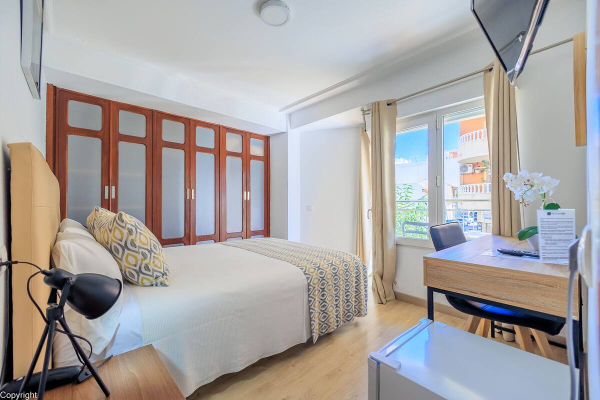 A hotel room with a bed, a desk with a chair, a lamp, and a window with curtains.