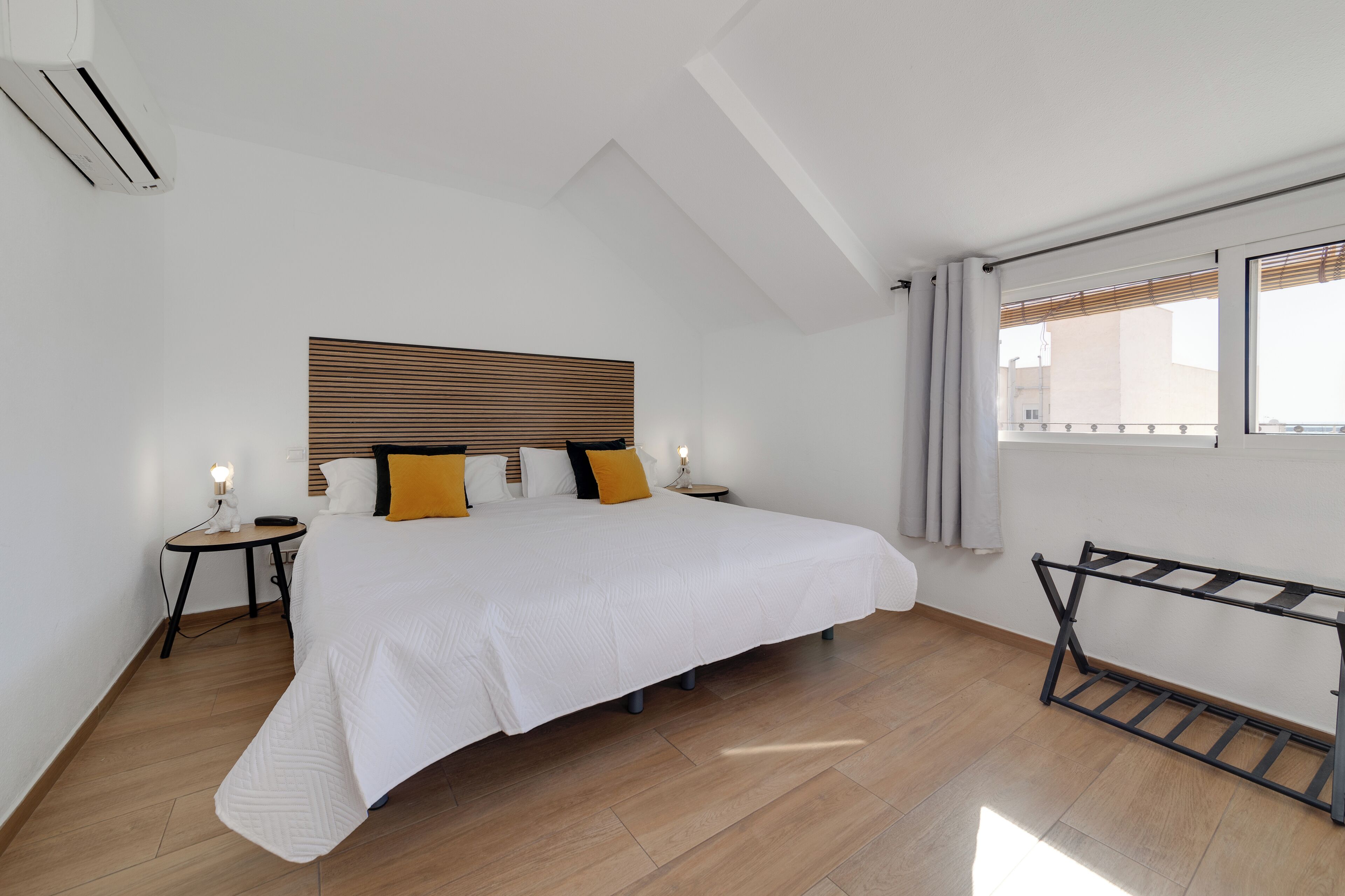 Une chambre avec un grand lit, une tête de lit en bois, une table de chevet avec une lampe, une fenêtre avec des stores et un porte-vêtements en métal.