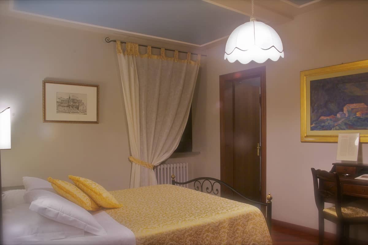 A bedroom with a bed, a desk, a chair, a radiator, a door, a window with curtains, and a painting on the wall.