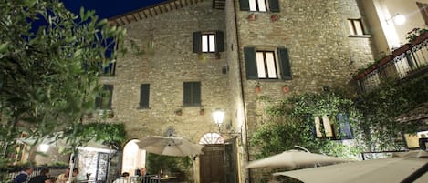 An outdoor cafe area with tables and umbrellas, surrounded by stone buildings with windows and a wooden door.