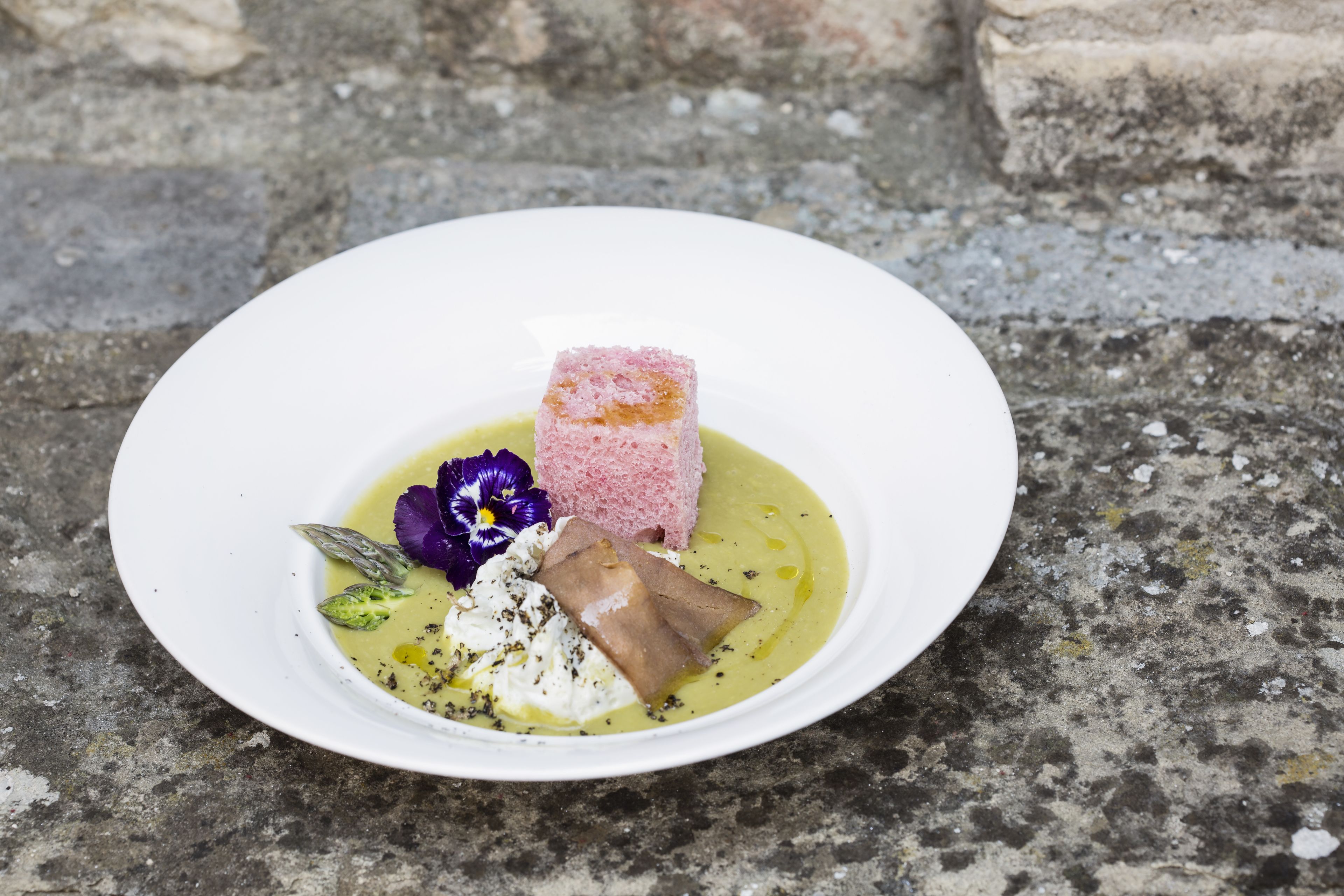 Een wit bord met een dessert bestaande uit een roze kubus, een stukje bruin vlees en een toef witte room, versierd met paarse bloemen, op een stenen oppervlak.