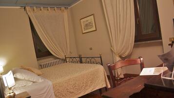 A bedroom with a bed, a nightstand, a chair, a desk, a radiator, and a window with curtains.