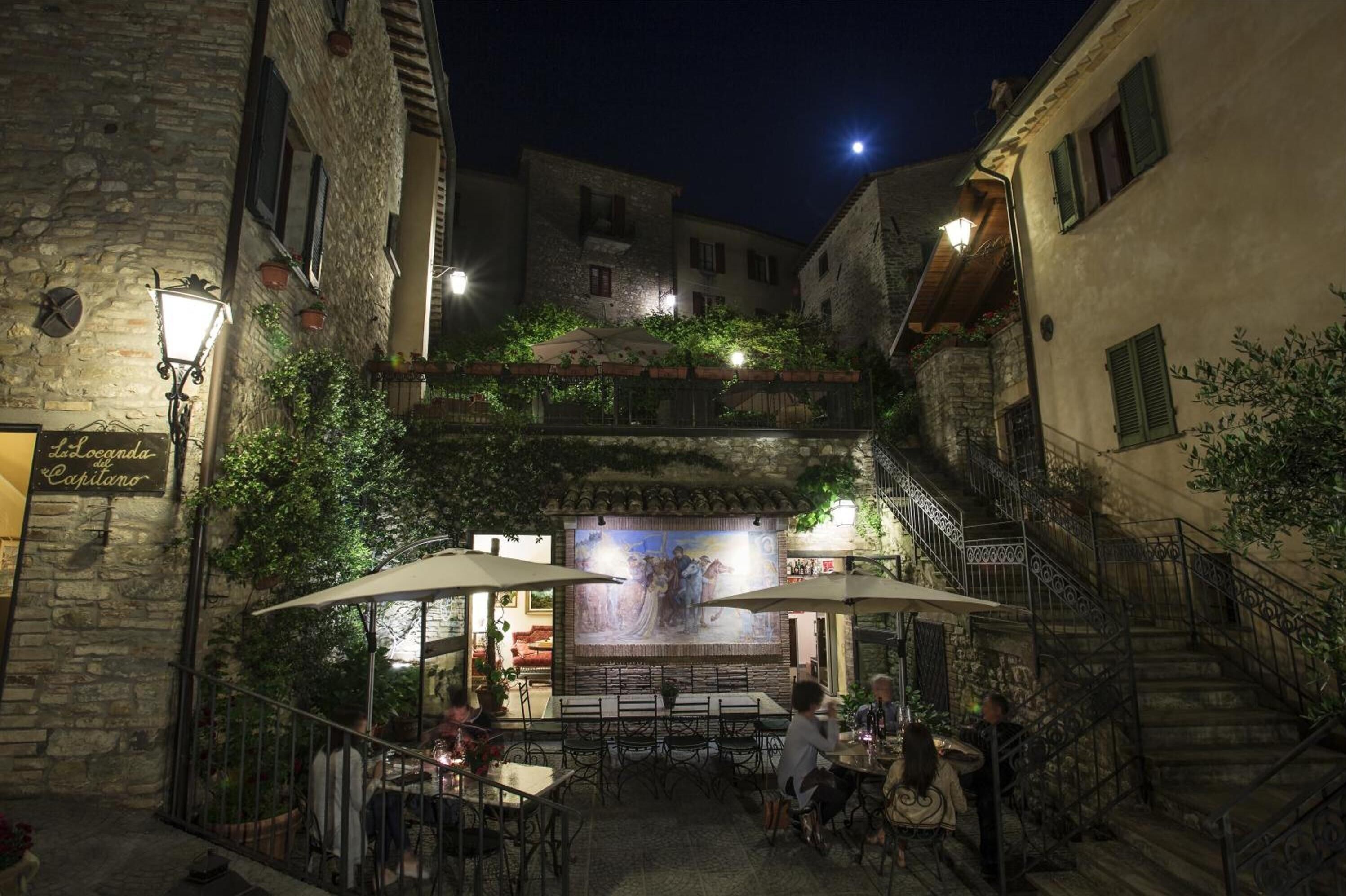 An outdoor café with stone buildings, outdoor seating, and a mural.