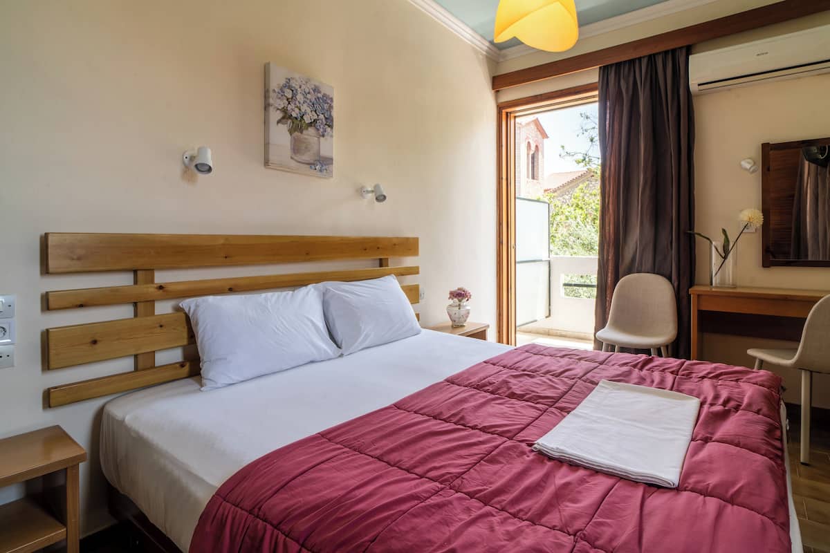 Un dormitorio con cabecera de madera, una cama grande con colcha roja, una mesita de noche, una silla y una ventana con vistas a un edificio y árboles.