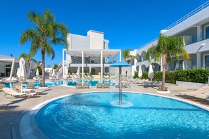 Una piscina con sillones de descanso y sombrillas, rodeada de palmeras y edificios modernos.