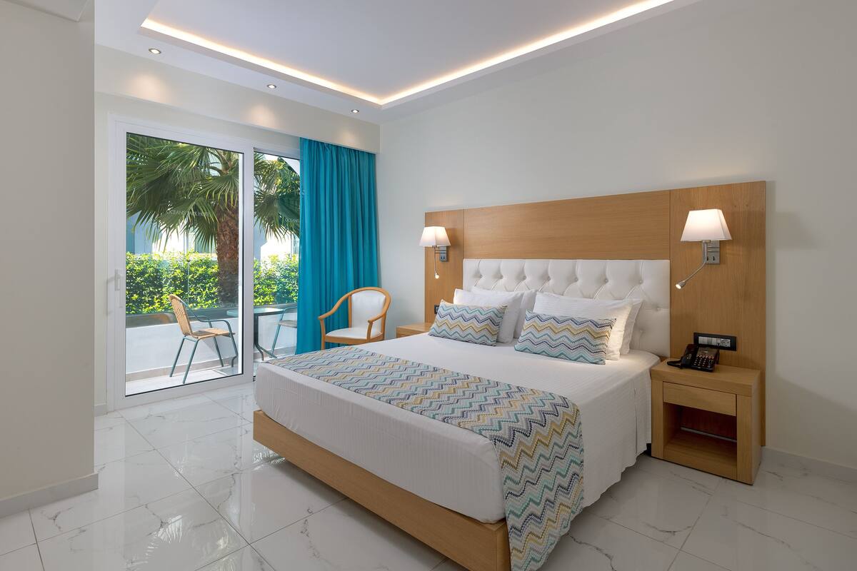 A modern hotel room with a large bed, two bedside lamps, a wooden nightstand, and a view of a palm tree through a sliding glass door.