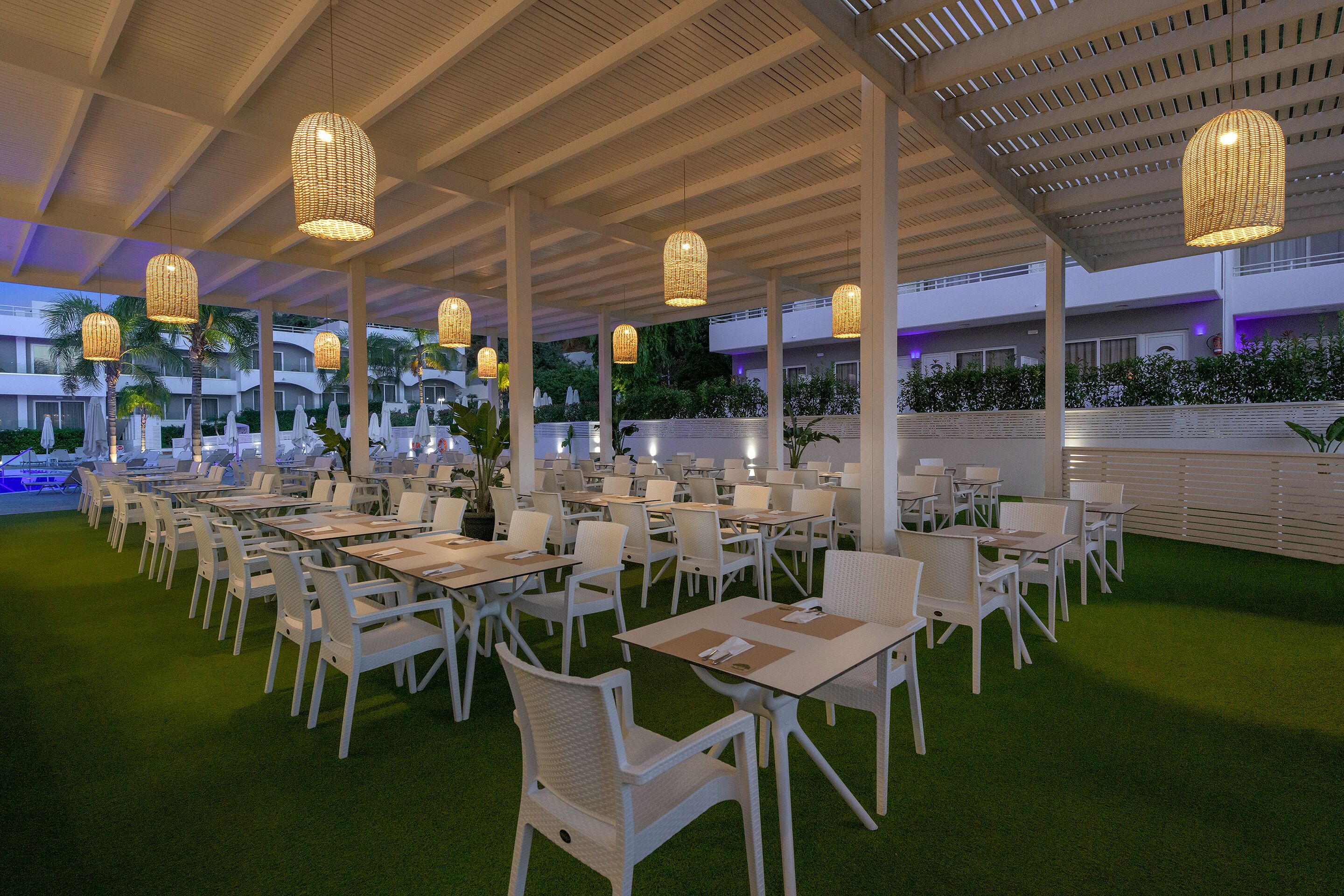 An outdoor dining area with white tables and chairs, hanging wicker lamps, and a green artificial turf floor.