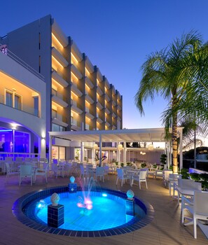 Un hotel moderno con zona de piscina, mobiliario exterior y fachada bien iluminada.