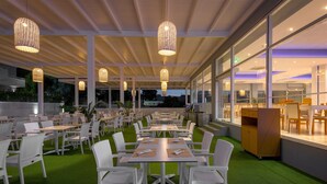 An outdoor dining area with white chairs and tables, green artificial turf, and hanging pendant lights. - Oceanis Park Hotel (Rhodes)
