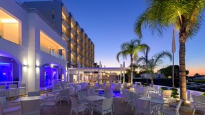 An outdoor dining area with white chairs and tables, a pool, and a modern building with balconies. - Oceanis Park Hotel (Rhodes)