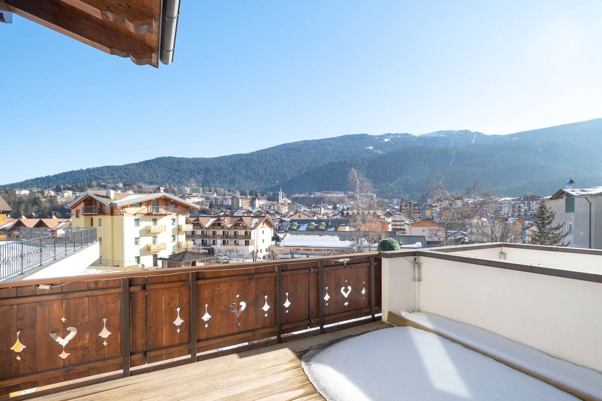 Un balcón con barandilla de madera y vistas a un pueblo con edificios y montañas al fondo.