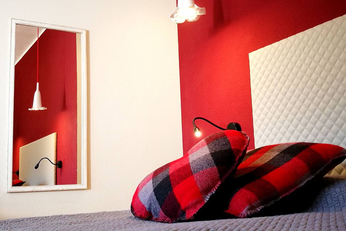 Un dormitorio con pared roja, cabecero blanco y espejo. Hay dos almohadas con estampado de cuadros y una lámpara de noche.