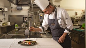 A chef in a professional kitchen with stainless steel surfaces, a large pot, and a plate of food. - Piccolo Hotel Andalo Suite Resort (Andalo)